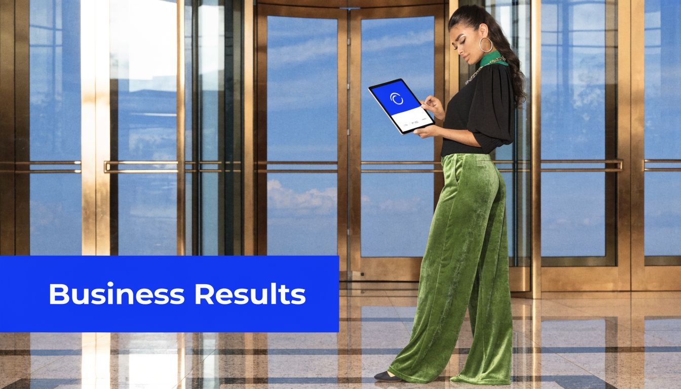 A businesswoman in green velvet pants stands in an office hallway holding a tablet showing corporate software.